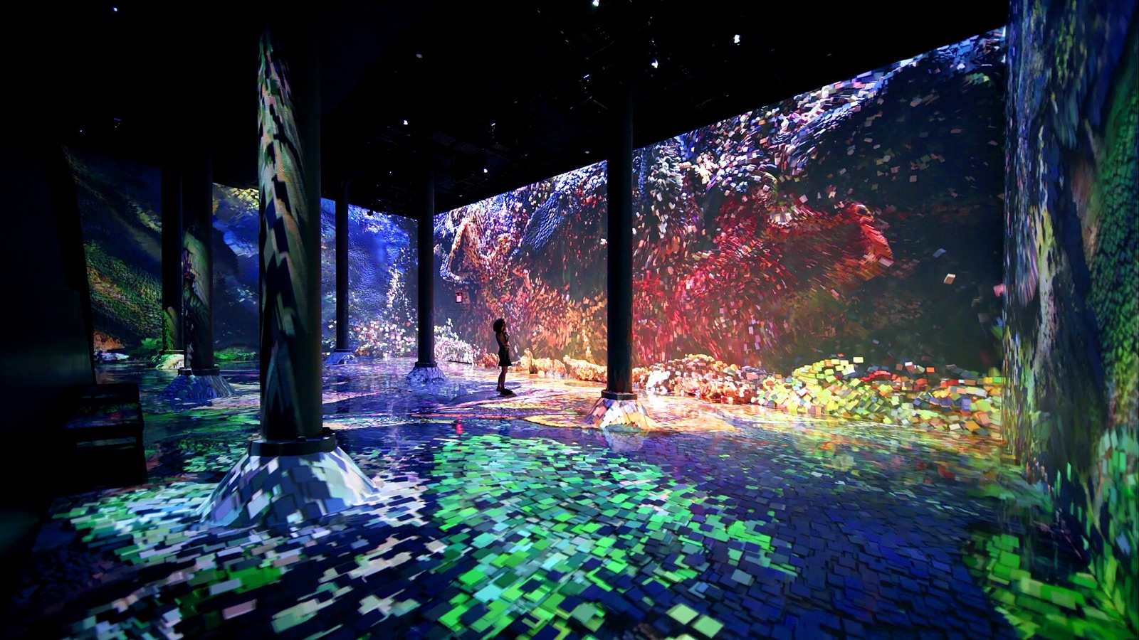 A dark hall with iron columns. Walls and floor are covered by projected squares of colour, red, green and blue, swirling like a coral reef; a single small figure stands in the middle looking up.