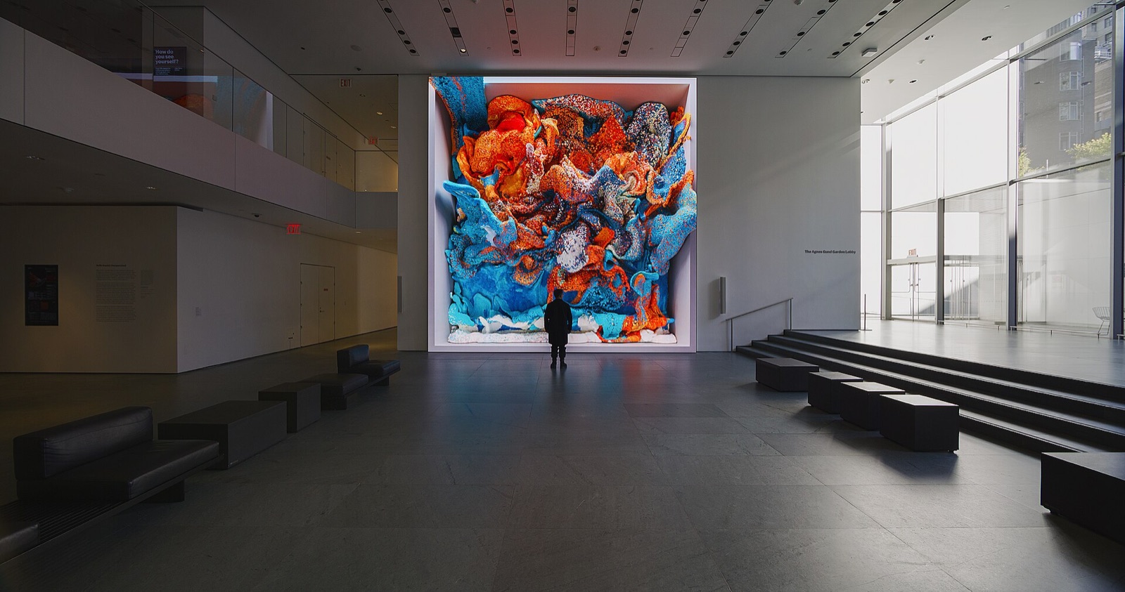 A dark museum lobby with black benches. On the far wall a screen as tall as two storeys shows a swirling mass of orange and turquoise shapes like coral or melted wax. A single person in a dark coat stands in front of it.