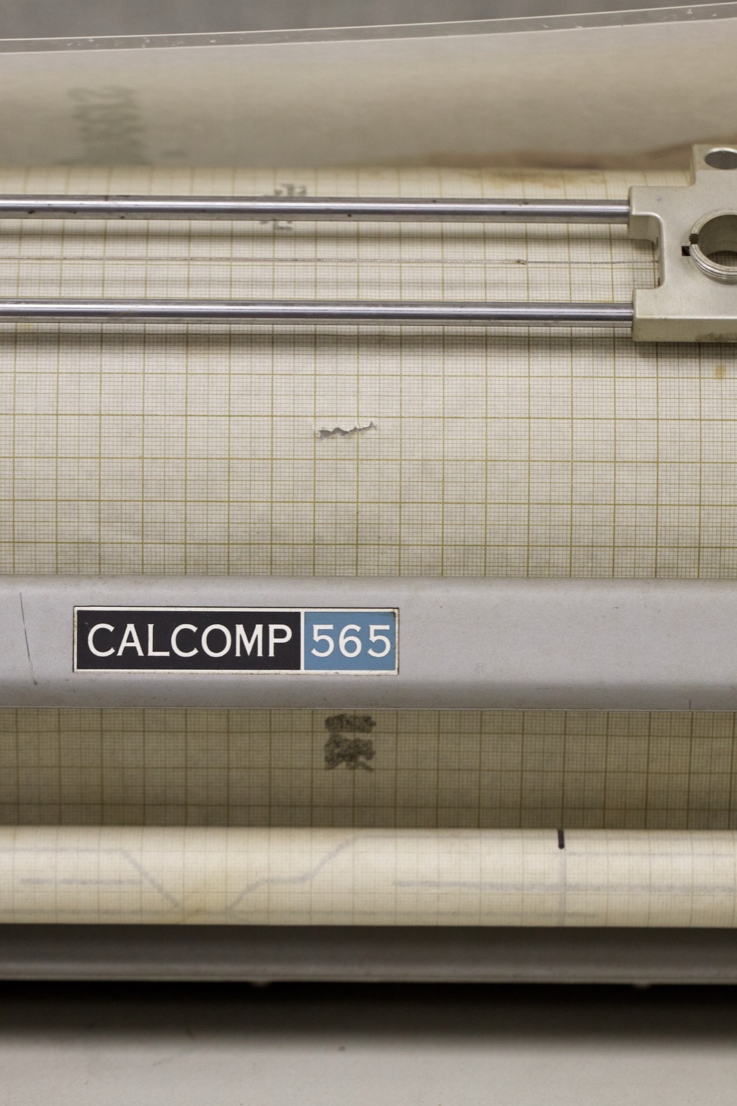 Close-up of a grey machine with the label CALCOMP 565. Above the label a sheet of graph paper lies over a drum, with two metal rods for the pen carriage.