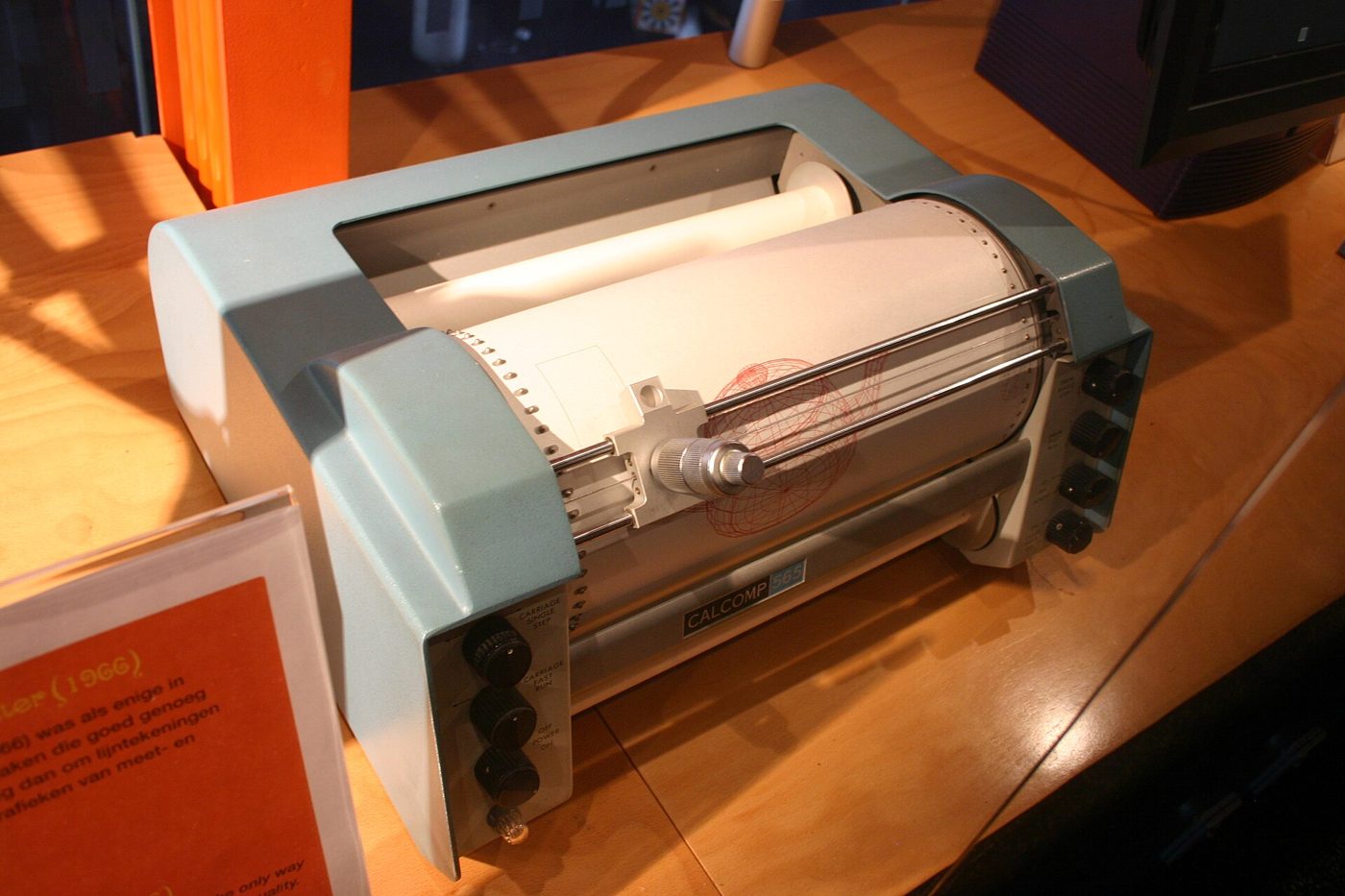 A pale blue drum plotter in a museum display: a roll of paper around a drum, with a pen carriage across it.