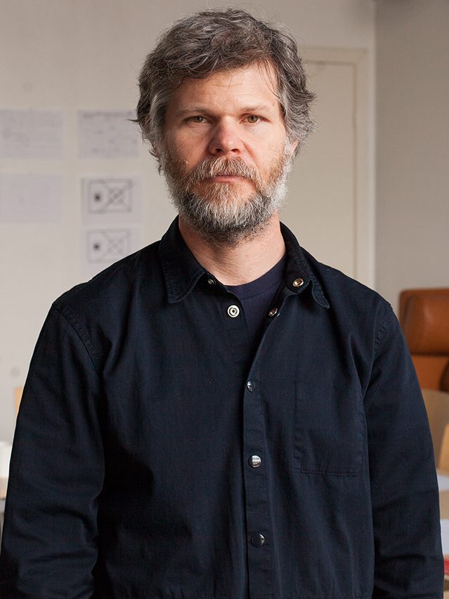 Casey Reas, a man with grey hair and a beard in a dark shirt, standing in a room with papers pinned to the wall and a brown sofa.