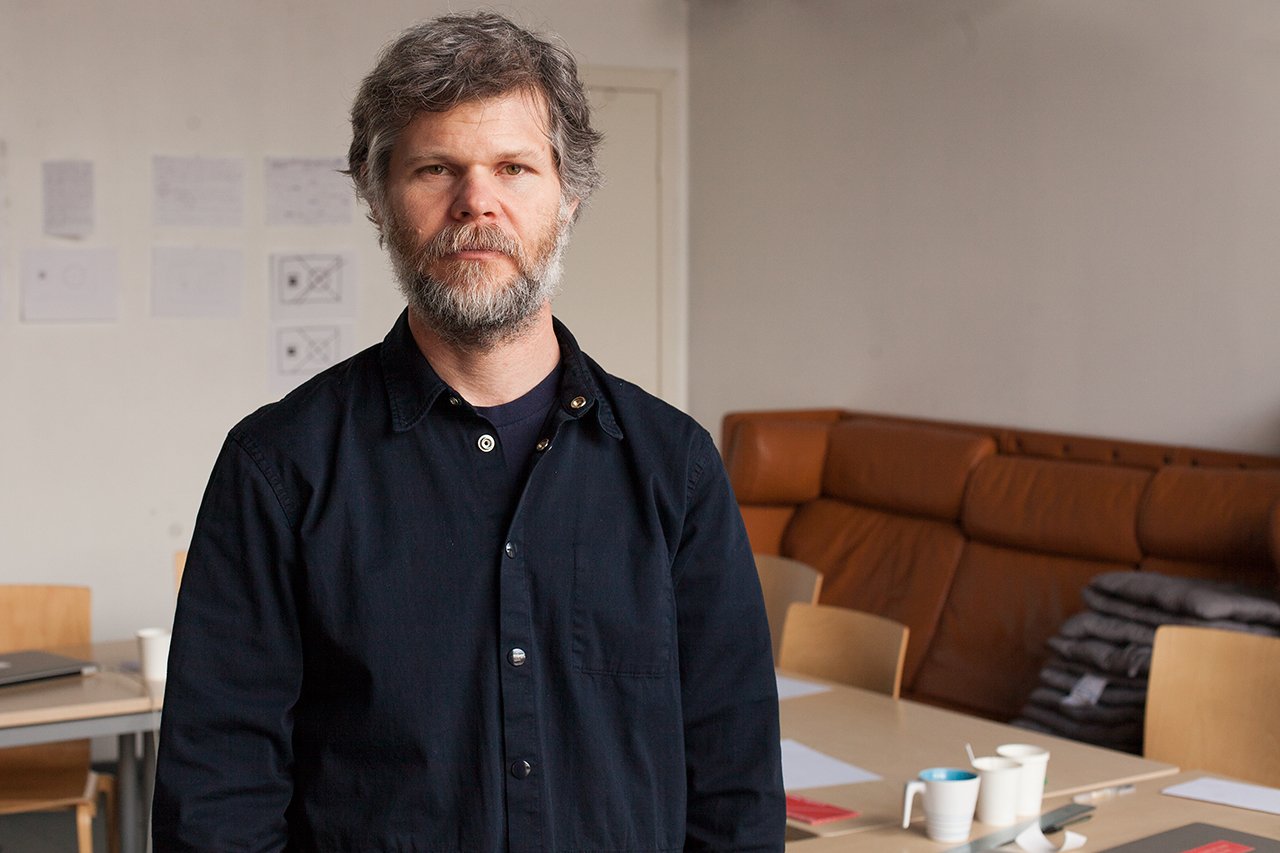 Casey Reas, a man with grey hair and a beard in a dark shirt, standing in a room with papers pinned to the wall and a brown sofa.