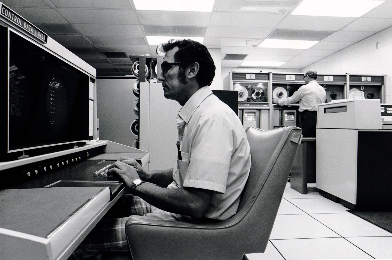 A black and white photograph of a 1960s computer room: a man in a short-sleeved shirt sits at a long grey console with a round screen, and behind him rows of tape drives and cabinets fill the room.