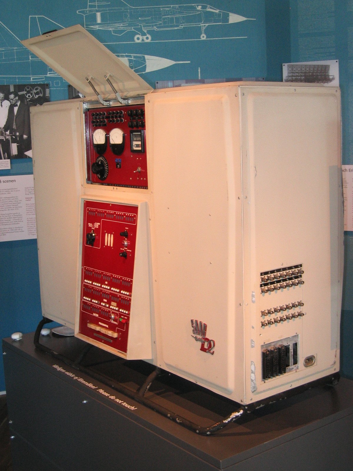 The Datasaab D2 in a museum: a cream-coloured cabinet on a low frame, with a red control panel full of switches, lamps and two round meters, and an open lid on top. An aircraft drawing hangs on the blue wall behind it.