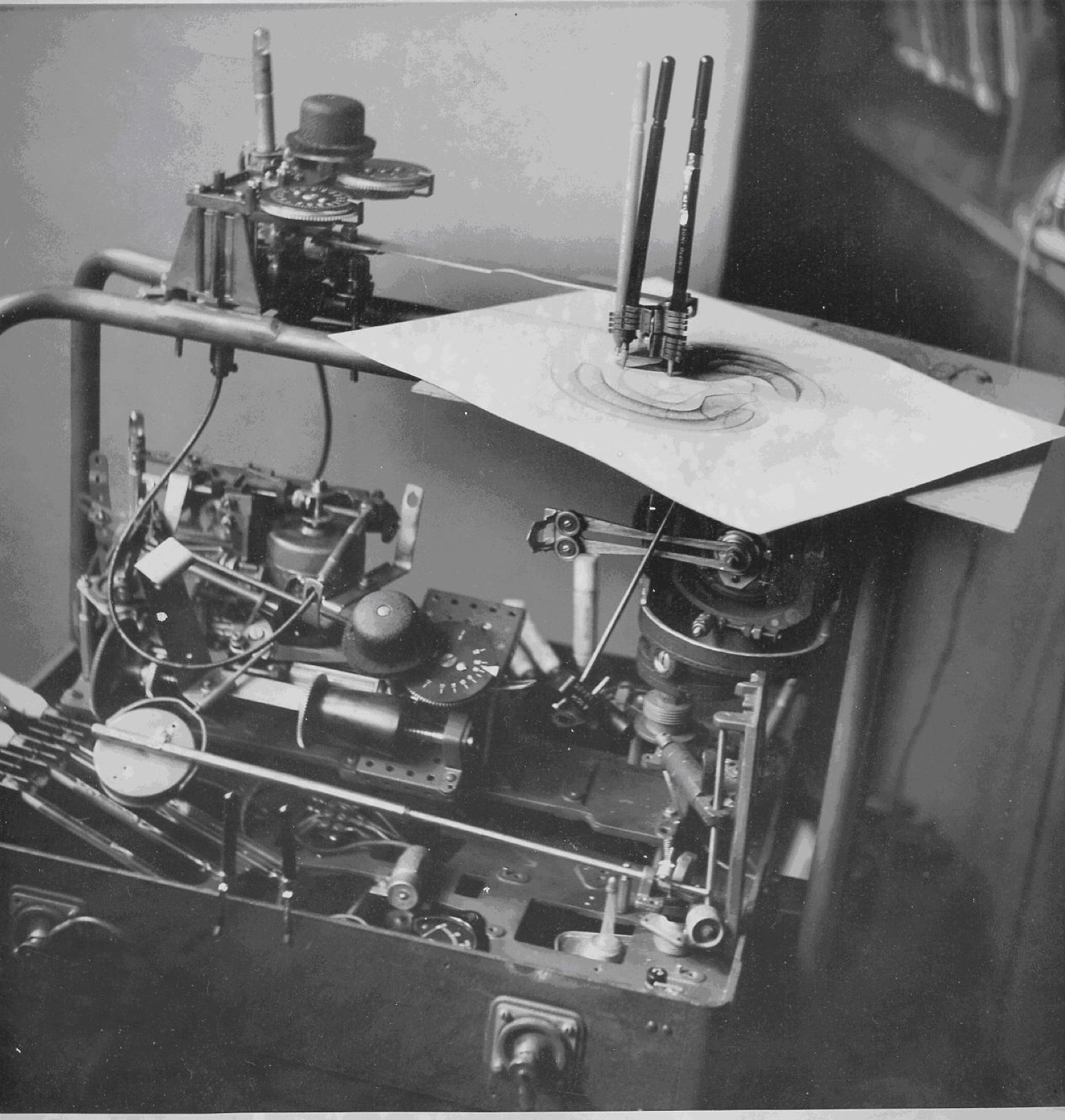 A black and white photograph of a dense mechanical device of gears, dials and rods. On top, three pens in a holder draw swirling curves on a sheet of paper.