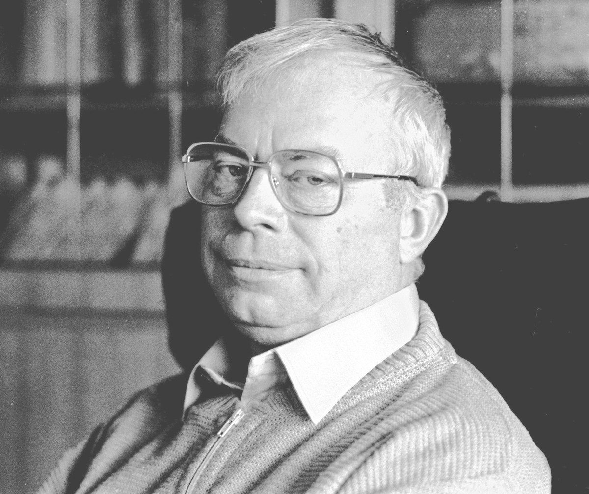 A black and white portrait of an older man with grey hair and glasses, in a light shirt and cardigan, looking calmly at the camera in front of a bookcase.