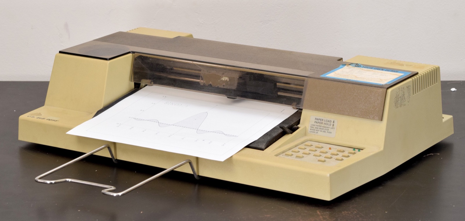 A beige desktop machine with a sheet of white paper loaded across its front and a row of buttons on the right.