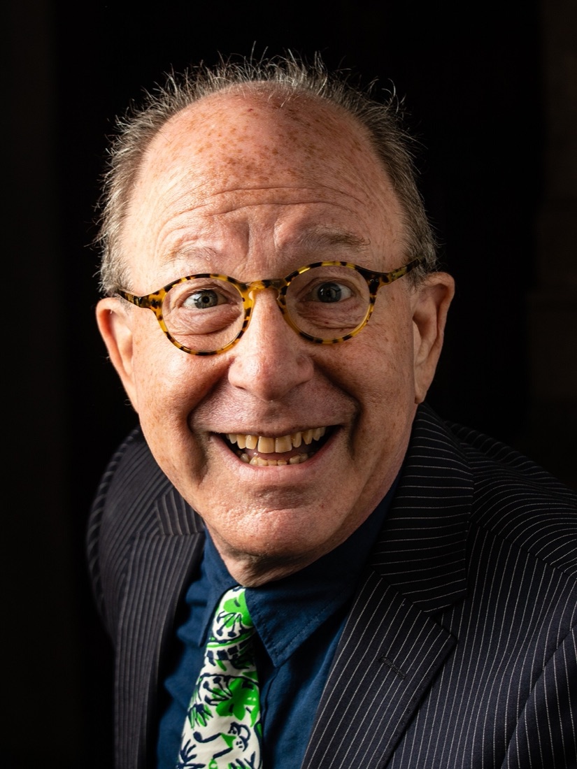 A smiling bald man with round tortoiseshell glasses, a pinstriped jacket and a green patterned tie, against a dark background.