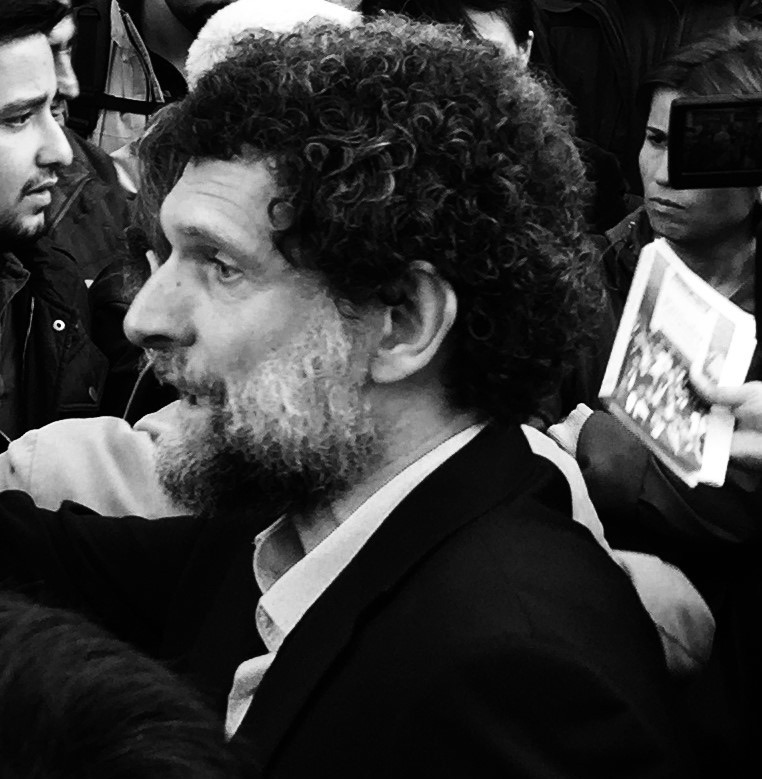 Black-and-white photo: a man with thick curly hair and a grey beard, in profile, in a dark jacket and open shirt, in a crowd; behind him someone holds up a leaflet.