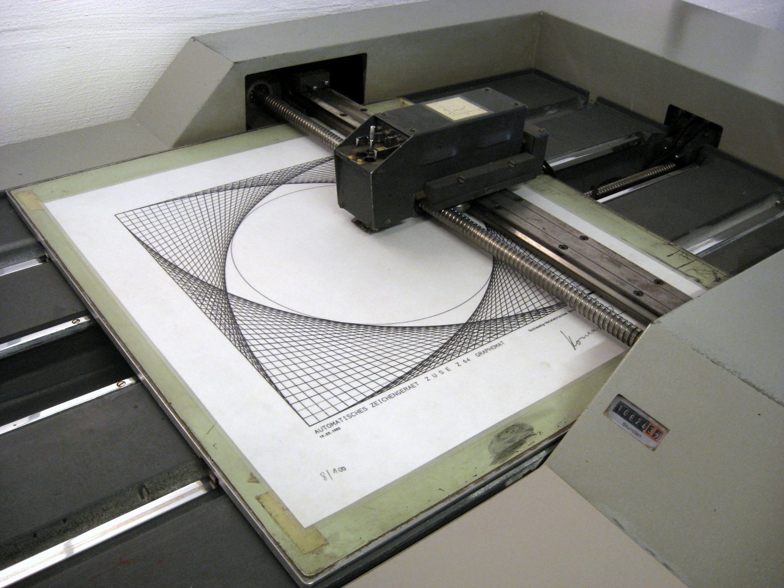 A flatbed drawing machine with a pen carriage on rails above a sheet of paper. The sheet shows a drawing of straight lines forming curves.