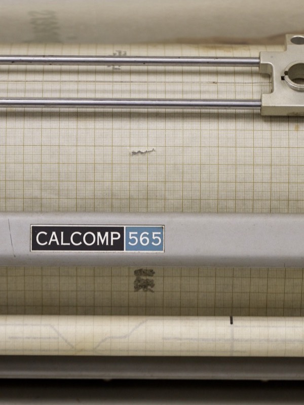 Close-up of a grey machine with the label CALCOMP 565. Above the label a sheet of graph paper lies over a drum, with two metal rods for the pen carriage; below, the paper roll with a faint plotted line.