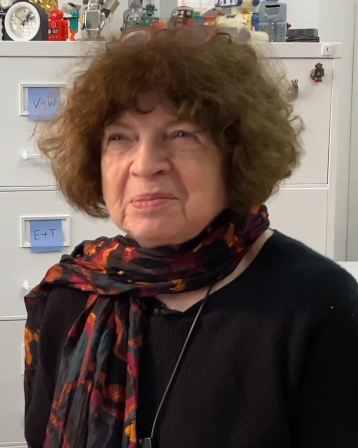 Jasia Reichardt, an older woman with curly brown hair, a dark sweater and a patterned red and black scarf, in front of grey filing cabinets with small toy robots on top.