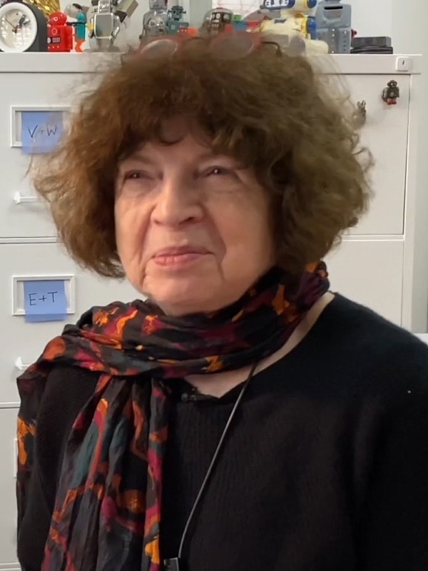 Jasia Reichardt, an older woman with curly brown hair, a dark sweater and a patterned red and black scarf, in front of grey filing cabinets with small toy robots on top.
