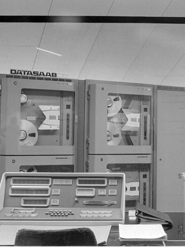 Black-and-white photo of a computer room: tall grey cabinets with magnetic tape reels behind glass, labelled DATASAAB D21, a desk console with rows of number displays and push-buttons in front, and an electric typewriter on the right.