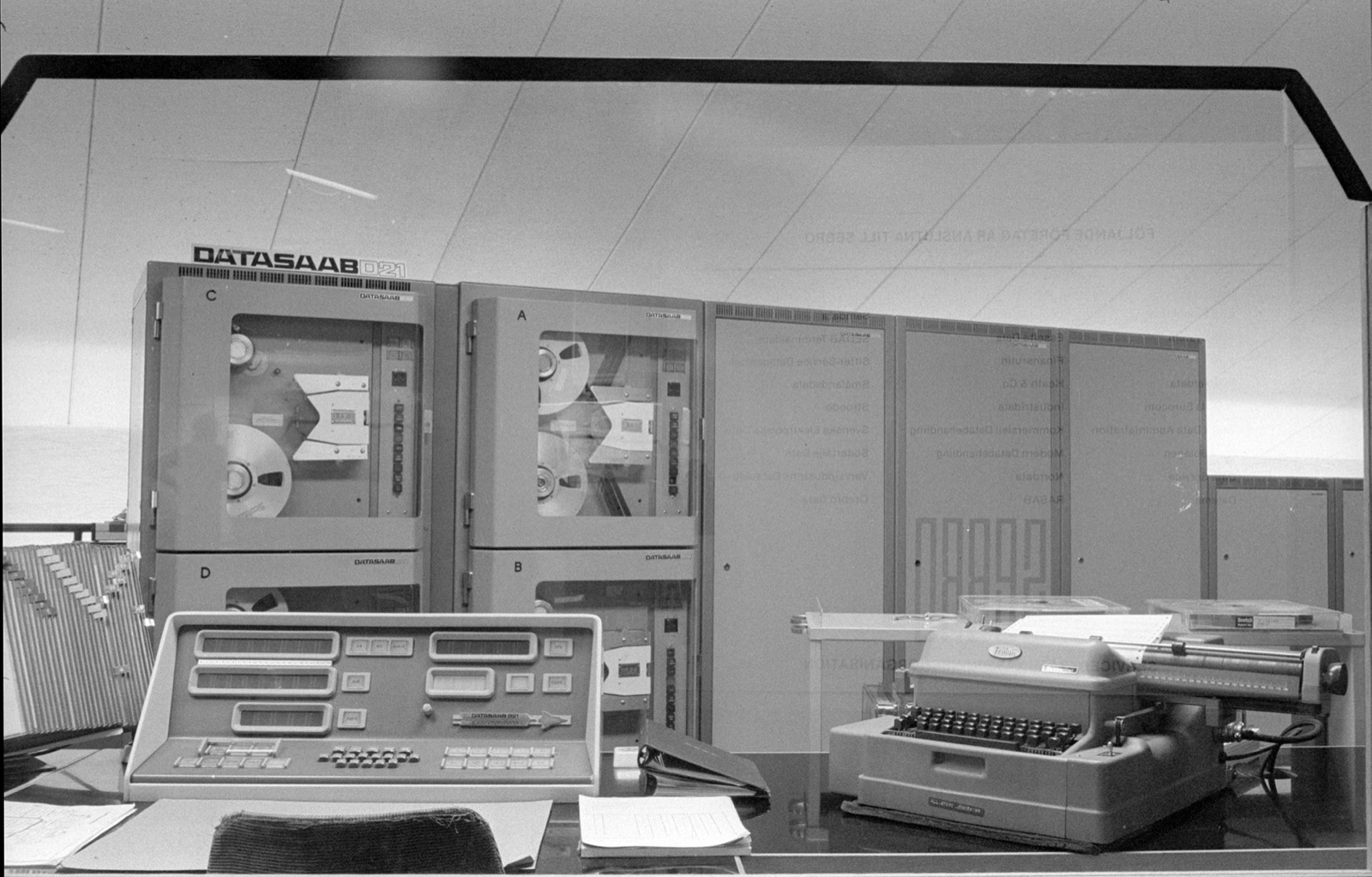 Black-and-white photo of a computer room: tall grey cabinets with magnetic tape reels behind glass, labelled DATASAAB D21, a desk console with rows of number displays and push-buttons in front, and an electric typewriter on the right.