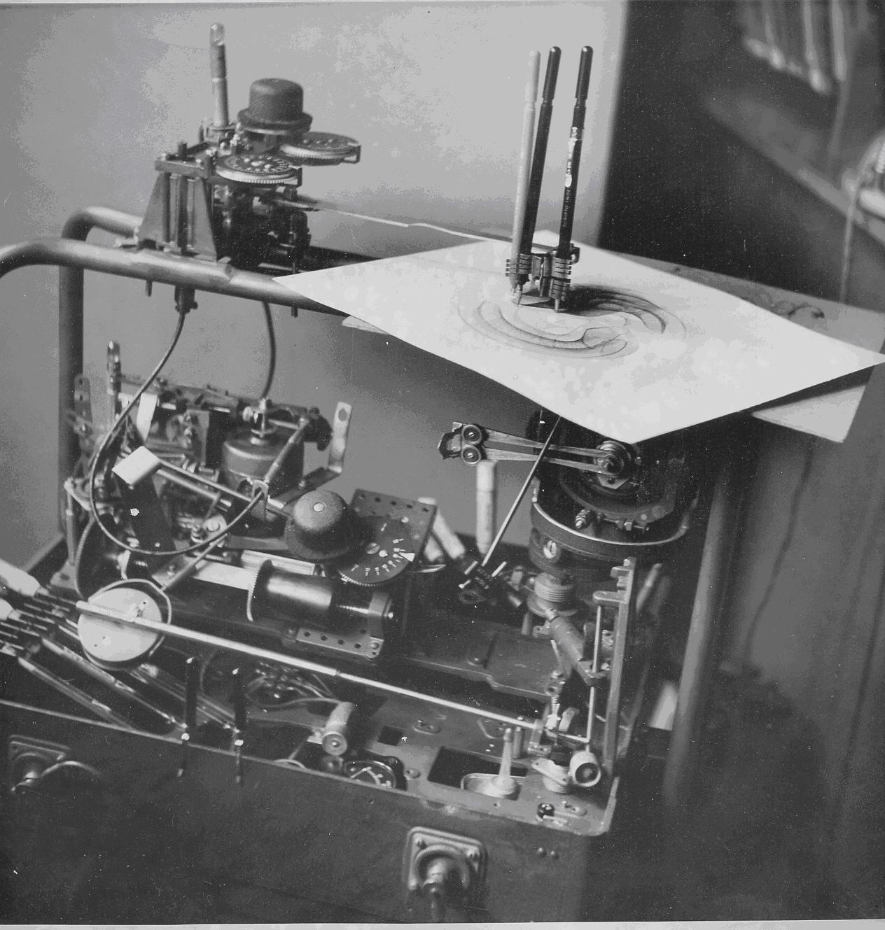 A black and white photograph of a dense mechanical device full of gears, dials, rods and cables. On top, three pens in a holder rest on a sheet of paper on which swirling curved lines have been drawn.