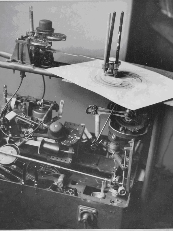 A black and white photograph of a dense mechanical device full of gears, dials, rods and cables. On top, three pens in a holder rest on a sheet of paper on which swirling curved lines have been drawn.