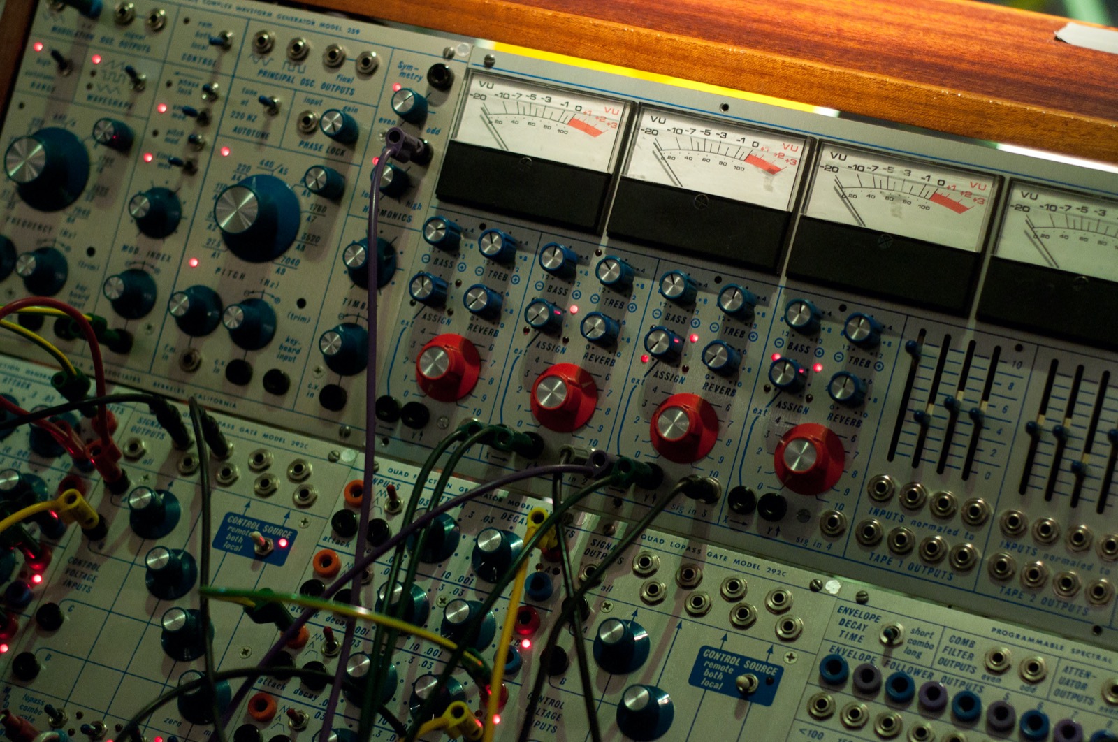 Close-up of a modular synthesizer: grey aluminium panels with blue and red knobs, four VU meters with needles, rows of sliders, and coloured patch cables plugged into small sockets.