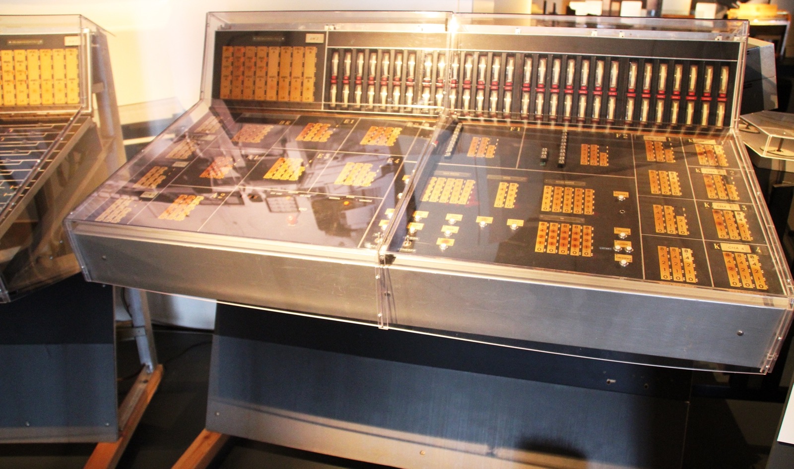 A wide control desk under clear plastic covers in a museum, with grids of orange push-buttons, a row of sliders along the back and labelled panels.