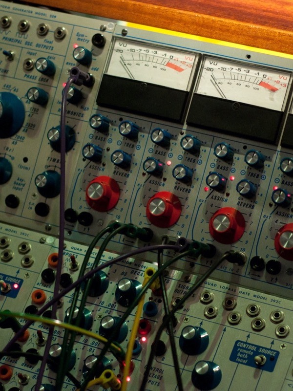 Close-up of a modular synthesizer: grey aluminium panels with blue and red knobs, four VU meters with needles, rows of sliders, and coloured patch cables plugged into small sockets.