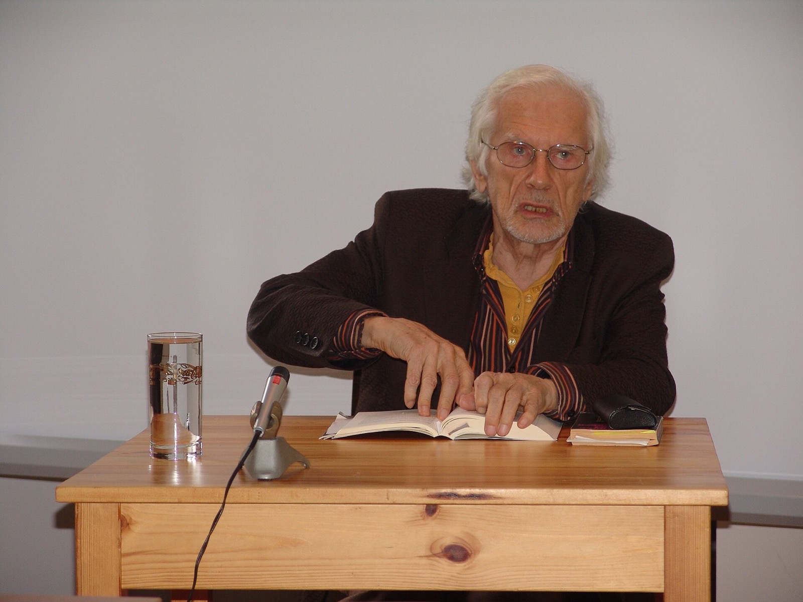 An elderly man with white hair and glasses sits at a pale wooden table in front of a white wall, speaking, with both hands resting on an open book. A microphone and a glass of water stand on the table.