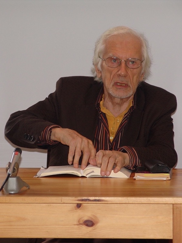An elderly man with white hair and glasses sits at a pale wooden table in front of a white wall, speaking, with both hands resting on an open book. A microphone and a glass of water stand on the table.