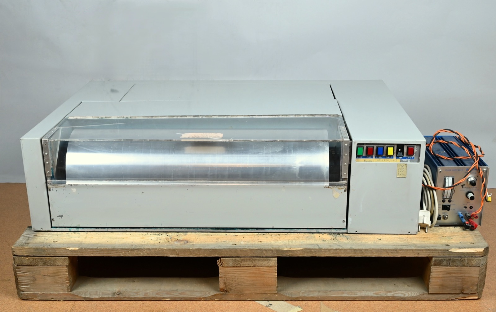 A grey metal machine on a wooden pallet: a long drum lies under a clear plastic cover, with a control panel of green, red, blue and yellow buttons on the right and a blue power supply with orange cables beside it.