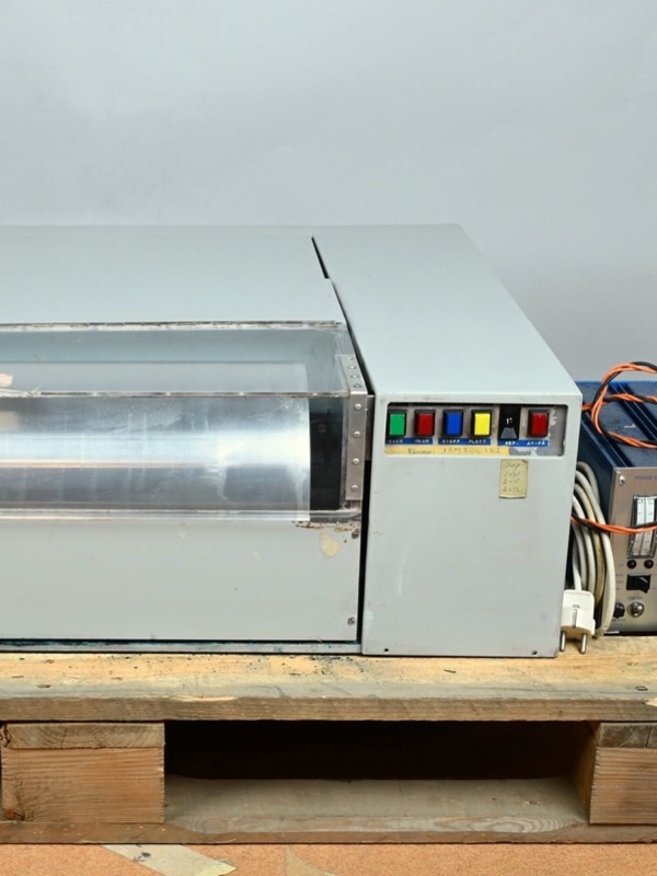 A grey metal machine on a wooden pallet: a long drum lies under a clear plastic cover, with a control panel of green, red, blue and yellow buttons on the right and a blue power supply with orange cables beside it.
