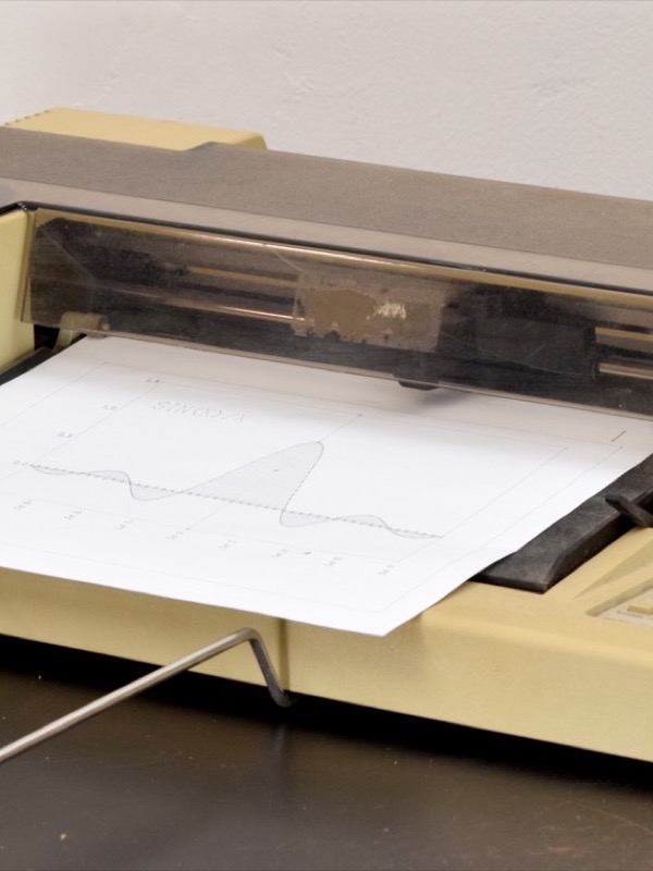 A beige desktop machine seen from the front. A sheet of white paper with a plotted graph of a wave lies across it under a smoked plastic cover with a metal rail and the pen carriage. A wire paper support sticks out in front, and a panel of small buttons sits on the right.
