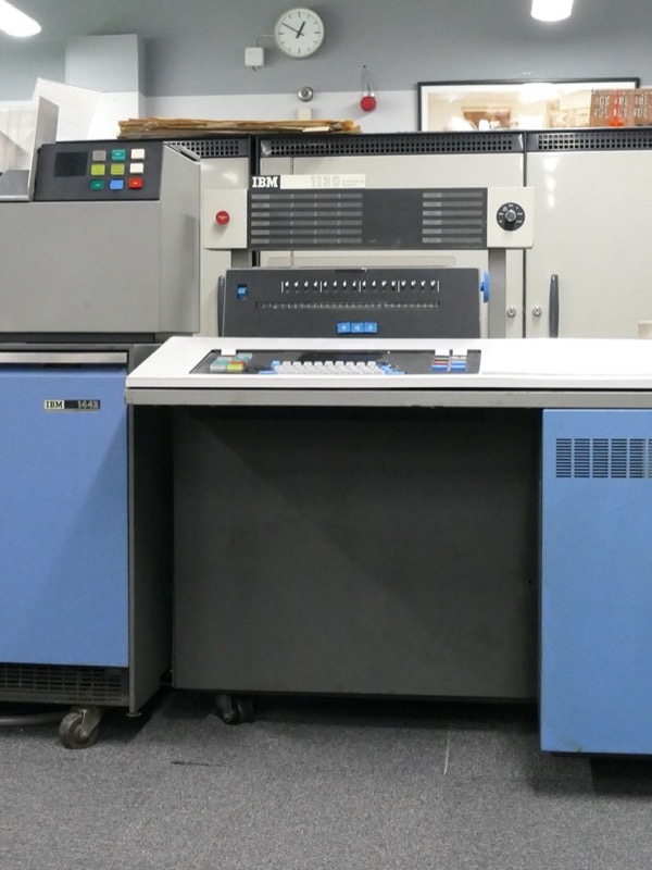 An IBM 1130 system in a museum: a grey card reader on blue cabinets on the left, and on the right the central unit, a white desk with a panel of indicator lights, a keyboard and a blue cabinet.