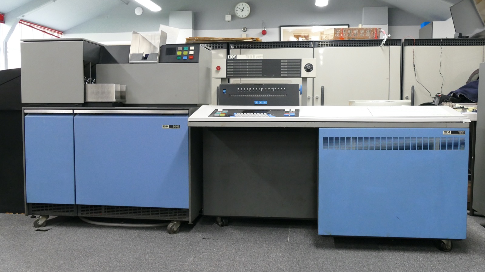An IBM 1130 system in a museum: a grey card reader on blue cabinets on the left, and on the right the central unit, a white desk with a panel of indicator lights, a keyboard and a blue cabinet.