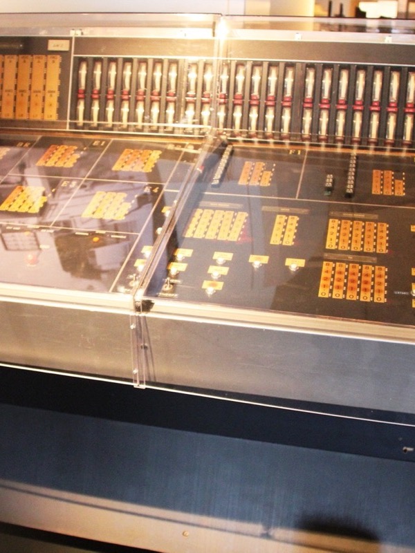A wide control desk under clear plastic covers in a museum, with grids of orange push-buttons, a row of sliders along the back and labelled panels.