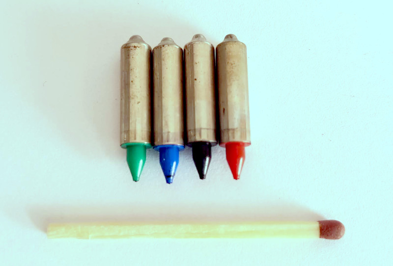 Four short metal pen cartridges standing in a row on a white surface, each with a coloured conical tip: green, blue, black and red. Below them lies a wooden match with a brown head, a little longer than the four pens are wide together.