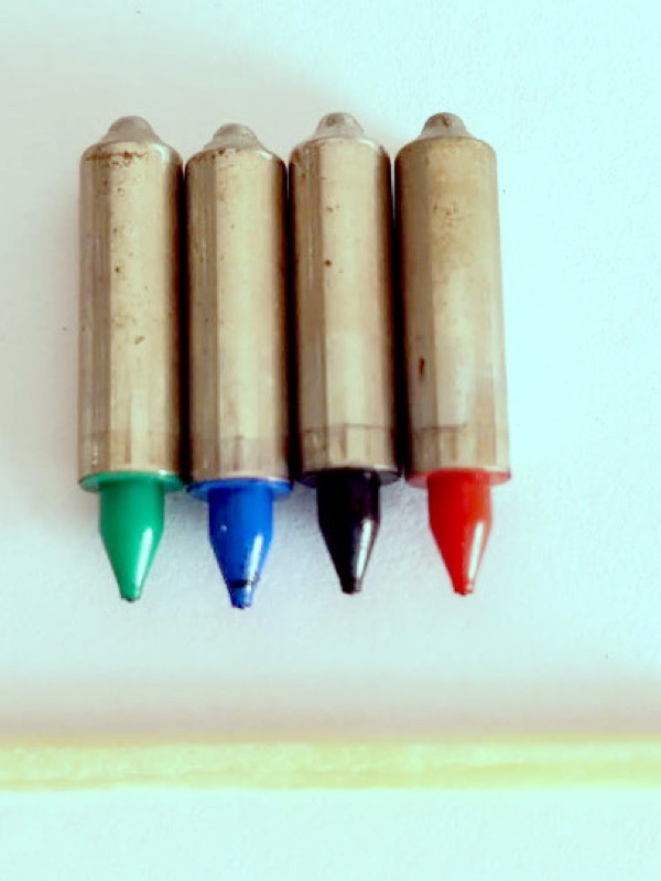 Four short metal pen cartridges standing in a row on a white surface, each with a coloured conical tip: green, blue, black and red. Below them lies a wooden match with a brown head, a little longer than the four pens are wide together.