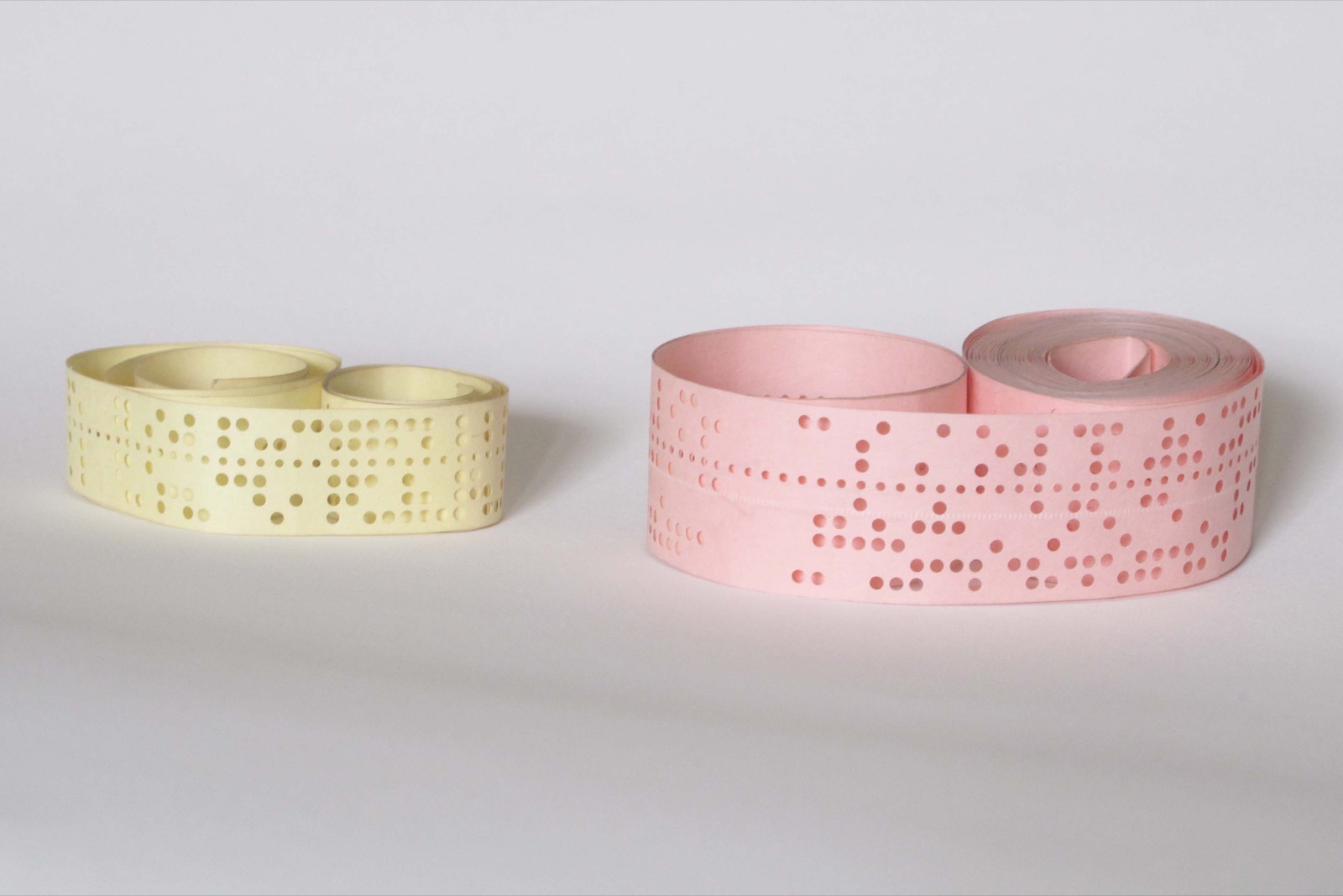 Two loose rolls of punched paper tape on a white surface: a narrower cream-coloured one on the left and a wider pink one on the right. Both are punched with rows of round holes across the tape, with a line of smaller holes running along each.