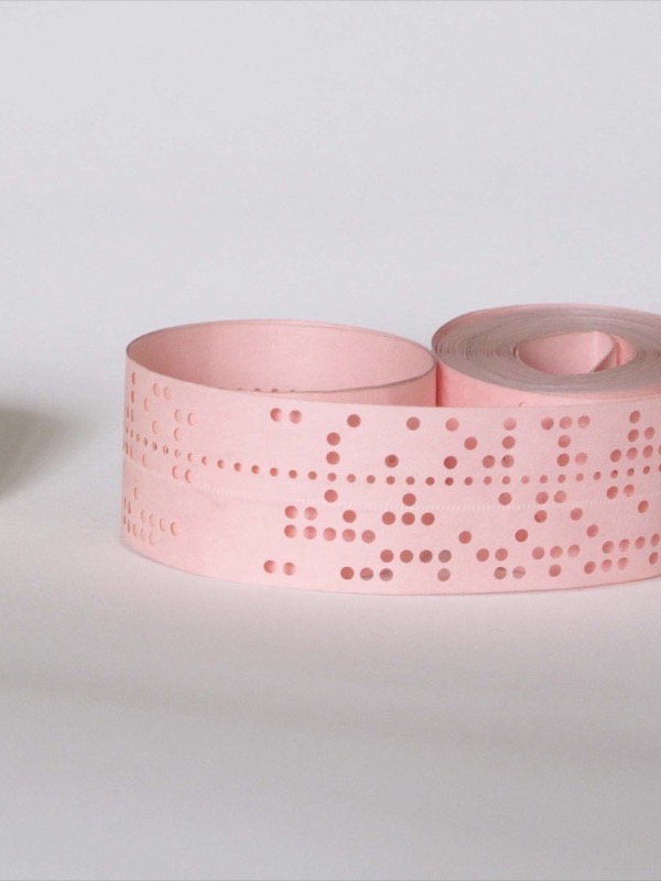 Two loose rolls of punched paper tape on a white surface: a narrower cream-coloured one on the left and a wider pink one on the right. Both are punched with rows of round holes across the tape, with a line of smaller holes running along each.