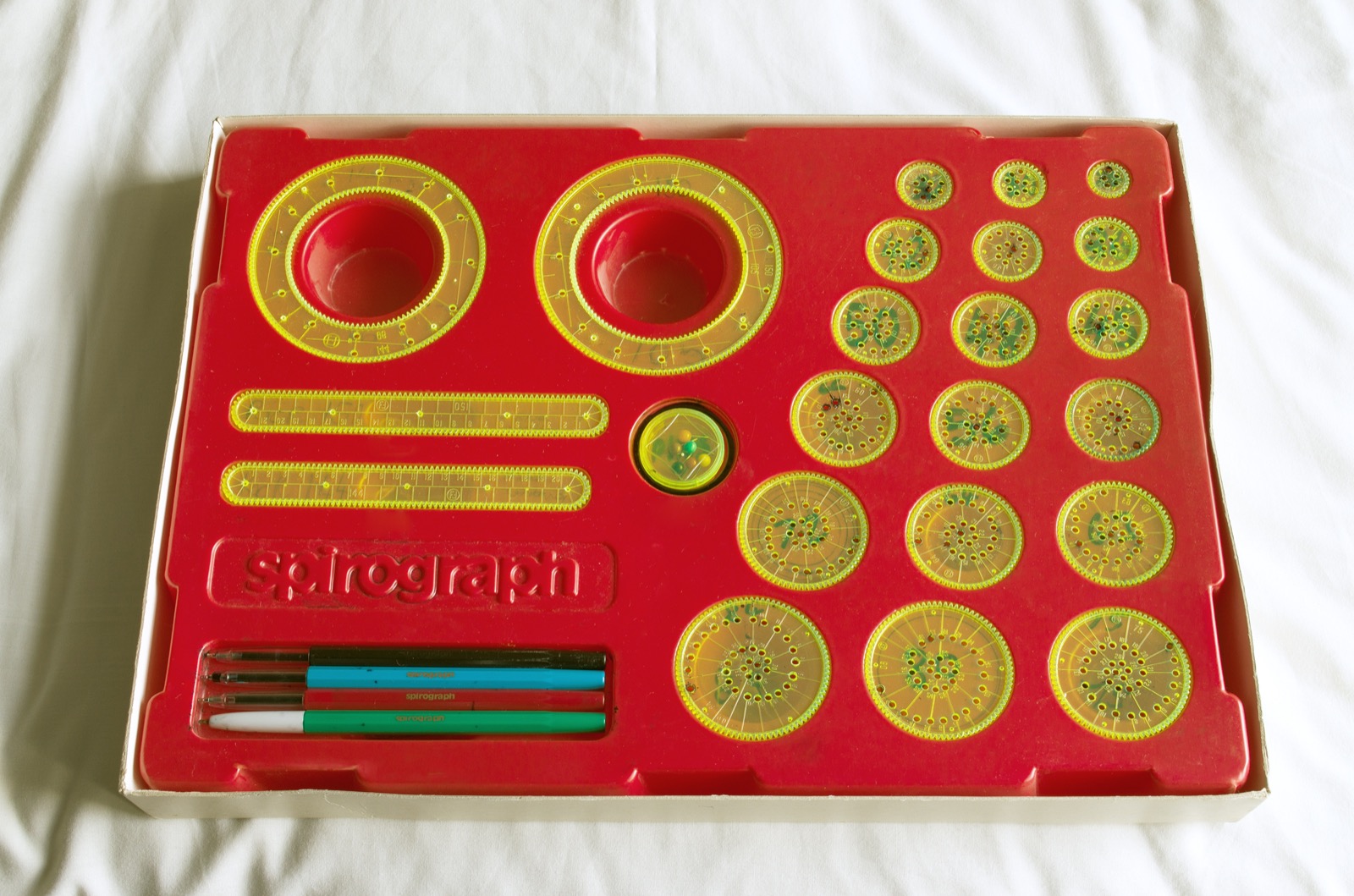 A Spirograph set in its red plastic tray, seen from above: two large transparent yellow rings with teeth inside and out, two toothed bars, eighteen round toothed wheels of different sizes full of small pen holes, and four ballpoint pens.