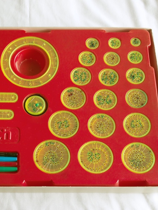 A Spirograph set in its red plastic tray, seen from above: two large transparent yellow rings with teeth inside and out, two toothed bars, eighteen round toothed wheels of different sizes full of small pen holes, and four ballpoint pens.