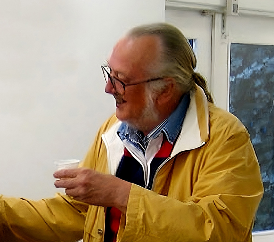 Sture Johannesson, an older man with glasses, a grey beard and long grey hair tied back, in a yellow jacket, smiling and holding a small paper cup.