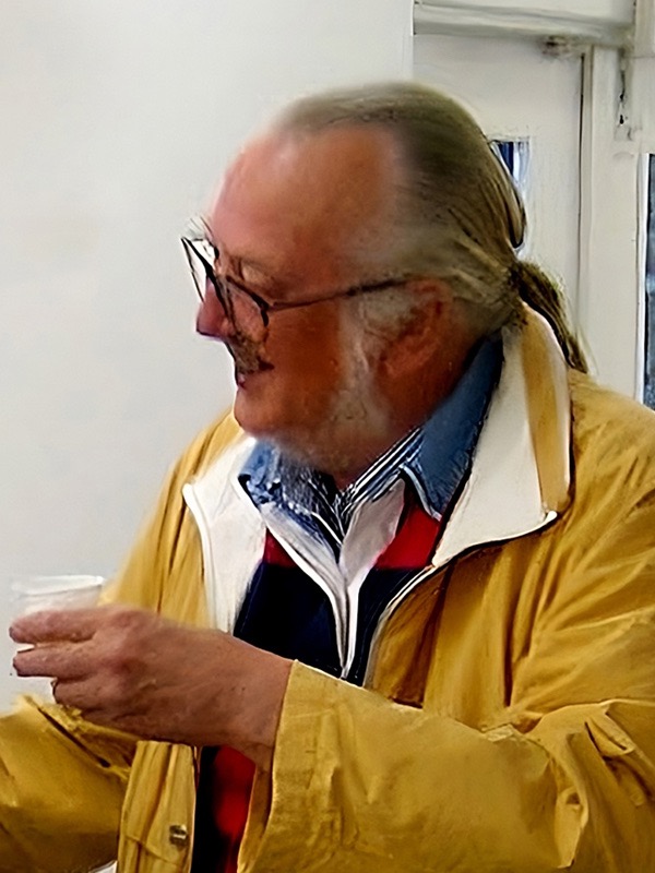 Sture Johannesson, an older man with glasses, a grey beard and long grey hair tied back, in a yellow jacket, smiling and holding a small paper cup.