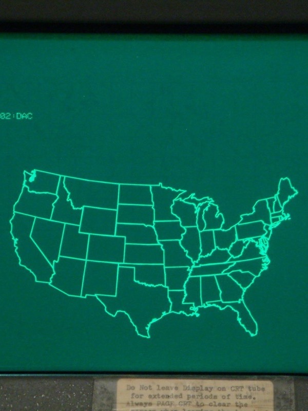 A green screen in a dark grey frame. Thin bright green lines draw the outline of the United States with the borders of every state. Top left, a few lines of green text. Below the screen a yellowed paper label begins: Do not leave display on CRT tube for extended periods of time. Always PAGE CRT to clear the screen.