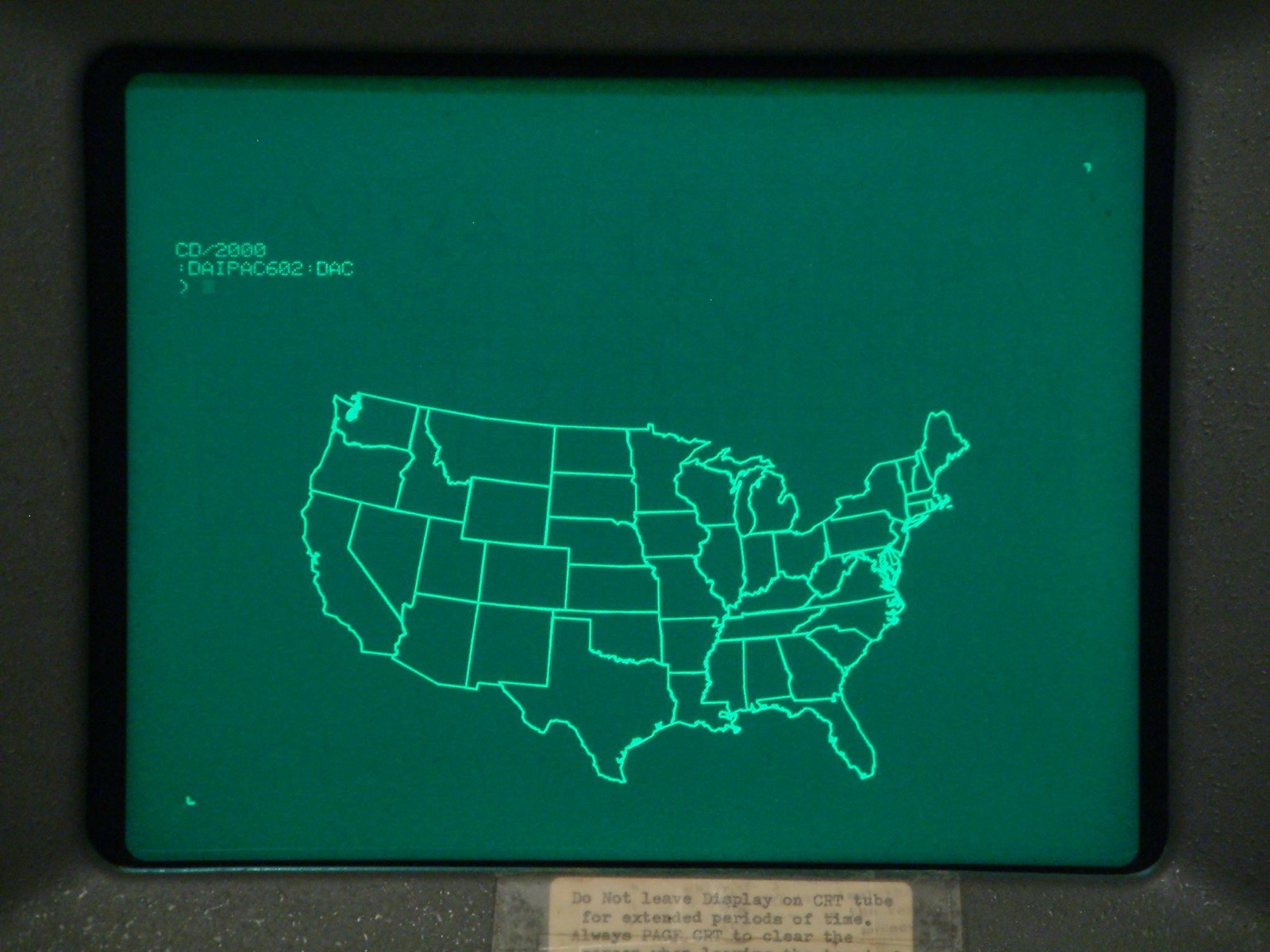 A green screen in a dark grey frame. Thin bright green lines draw the outline of the United States with the borders of every state. Top left, a few lines of green text. Below the screen a yellowed paper label begins: Do not leave display on CRT tube for extended periods of time. Always PAGE CRT to clear the screen.