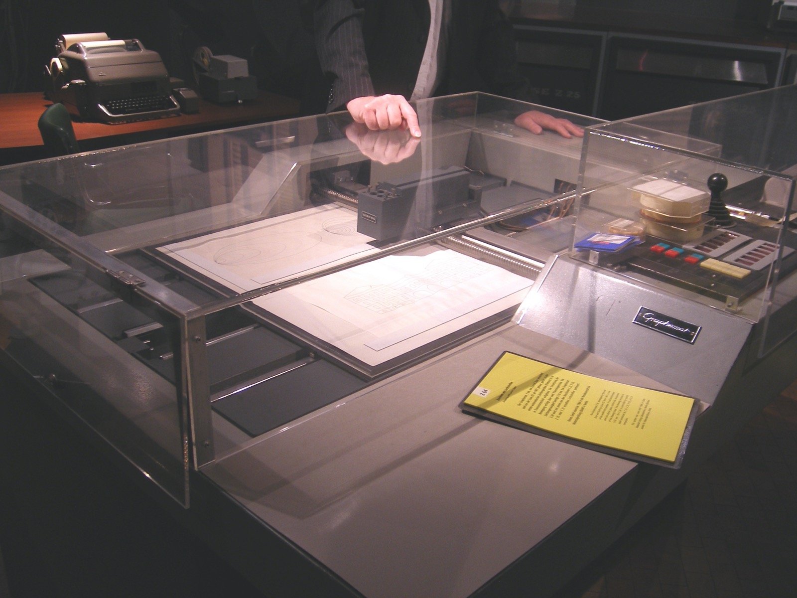 A large flatbed drawing machine under a clear plastic cover in a museum: a grey pen head on a rail above sheets of paper with line drawings of ellipses and a building facade, a control panel with buttons on the right, a plate reading Graphomat, and a yellow label in front. A visitor's hand rests on the cover.