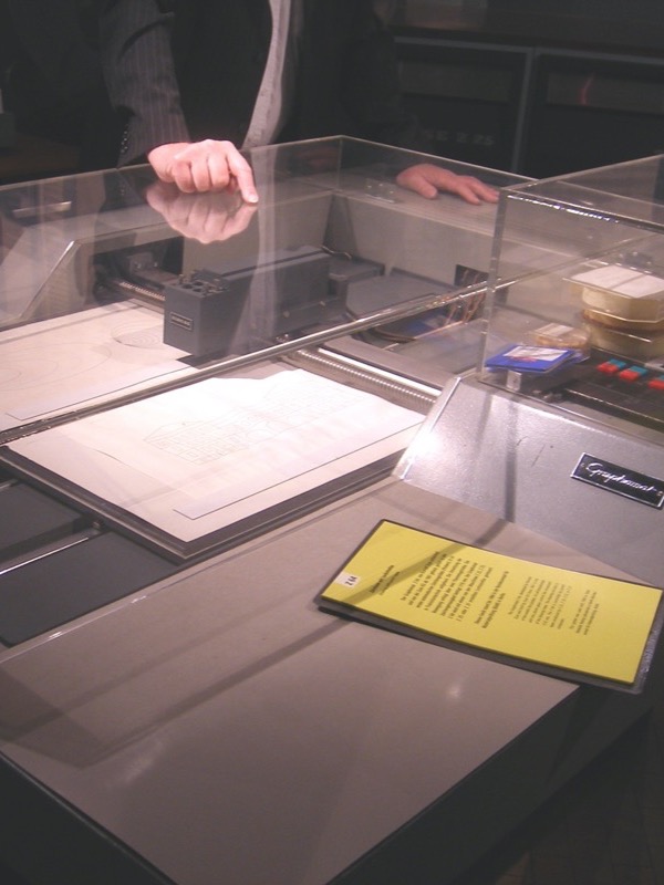 A large flatbed drawing machine under a clear plastic cover in a museum: a grey pen head on a rail above sheets of paper with line drawings of ellipses and a building facade, a control panel with buttons on the right, a plate reading Graphomat, and a yellow label in front. A visitor's hand rests on the cover.